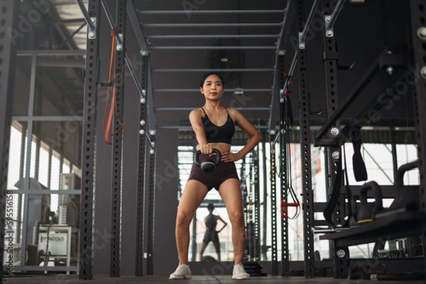 Fototapeta Strong asian woman doing exercise with kettlebell at crossfit gym. Athlete female wearing sportswear workout on grey gym background with weight and dumbbell equipment. Healthy lifestyle.