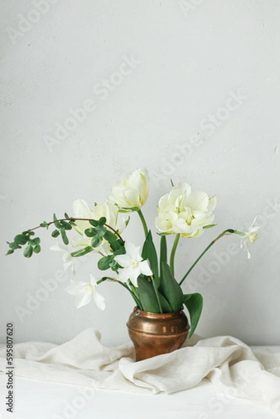 Fototapeta Happy Mothers day. Beautiful white tulips and daffodils in vintage vase on wooden table against rustic wall. Stylish simple spring bouquet, floral still life. Womens day. Space for text