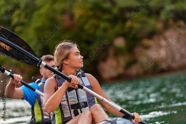 Fototapeta A young couple enjoying an idyllic kayak ride in the middle of a beautiful river surrounded by forest greenery