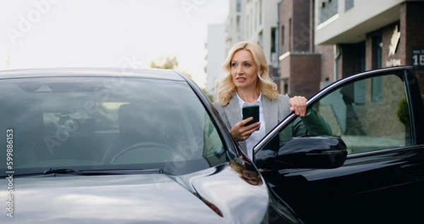 Fototapeta Good-looking positive and successful business 40-aged woman in formal clothes typing texting on she's mobile,standing near auto in outdoors on parking