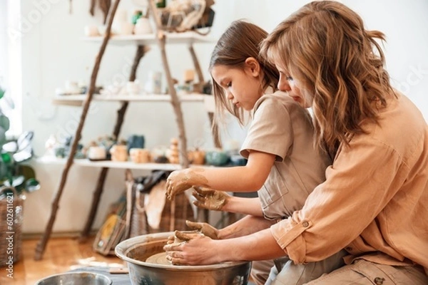 Fototapeta Pottery wheel. Mother with little girl making ceramic pot in the workshop