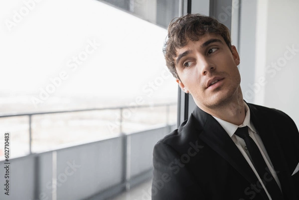 Fototapeta dreamy man in classic formal wear with black tie and white shirt standing in modern hotel room and looking at window, groom on wedding day, special occasion