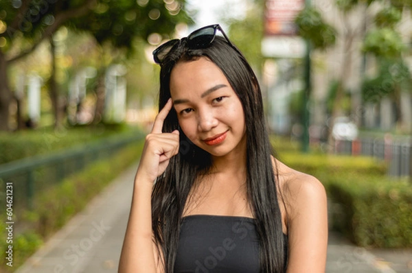 Fototapeta A pretty lady standing in the park wearing a black strapless tank top and sunglasses in a thinking pose smiling while standing outside, in the park. A trees, fence and bushes in the background.