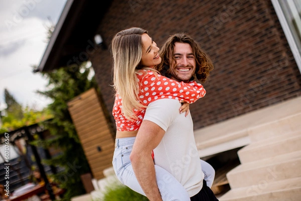 Fototapeta Handsome long hair man carrying the young woman on his back in front of brick house