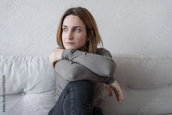 Fototapeta Young depressed woman sitting on sofa in living room. She feeling sad and worried suffering depression in mental health.