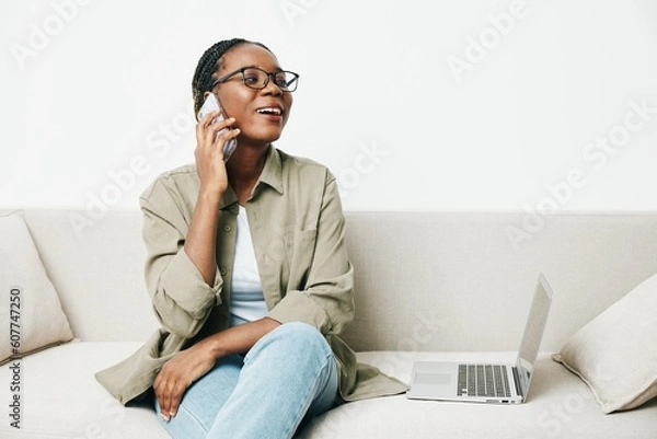 Fototapeta African American woman business freelancer working sitting on the couch at home in a laptop and phone, business calls and correspondence sadness and anger, home clothes and eyeglasses, light interior