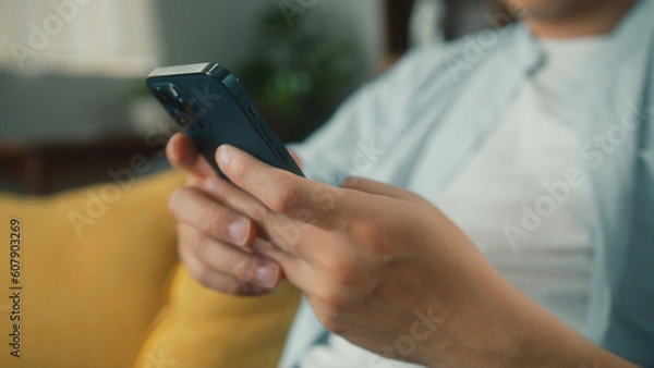 Fototapeta Closeup of Happy attractive Asian man sit on sofa hand holding mobile phone chatting and playing social media. Young male looking at smartphone cellphone browsing internet on sofa at home living room