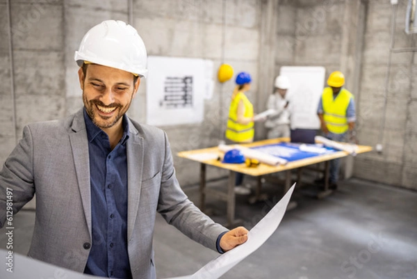 Fototapeta Civil engineer looking at building plans and blueprint at construction site while architects brainstorming in background.