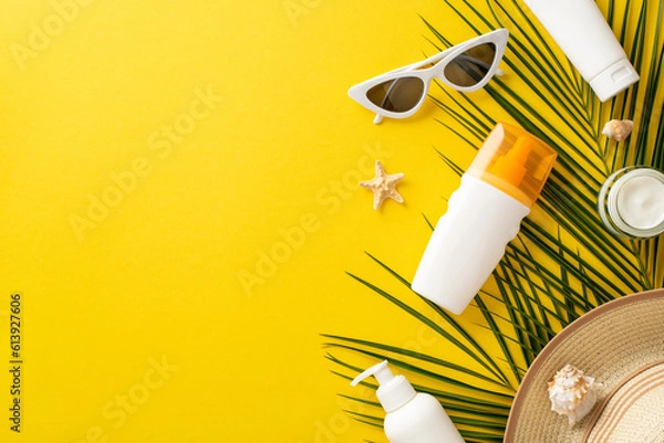 Fototapeta Relaxing under sun theme. Top view arrangement showcasing sunscreen bottles without labels, sunglasses, sunhat, seashells, starfish, palm leaf on bright yellow background with empty space for branding