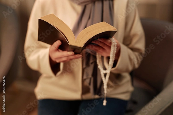 Fototapeta Focus on open quran in soft black cover held by young Muslim woman reading surahs consisting of verses and using rosary during prayer