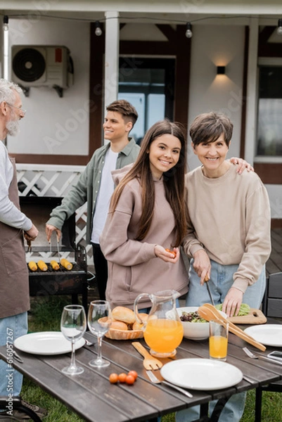 Fototapeta celebrating parents day, modern parenting, middle aged woman cutting lettuce near happy teenage daughter making salad, father and son grilling corn on bbq grill, on blurred background, bbq party