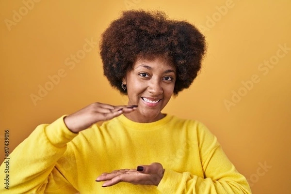 Fototapeta Young african american woman standing over yellow background gesturing with hands showing big and large size sign, measure symbol. smiling looking at the camera. measuring concept.