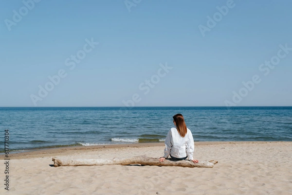 Fototapeta Lonely young woman in a white jacket sitting alone on a log on beach by sea, mood. Rear view of a woman in casual clothes alone on shore, copy space