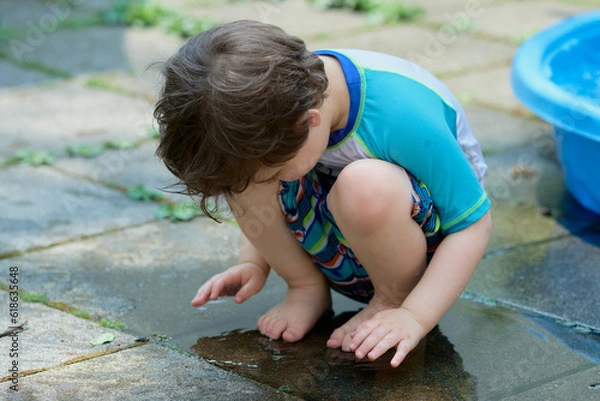 Fototapeta young boy in a bathing suit playing in a puddle of water in the backyard next to a shallow swimming pool