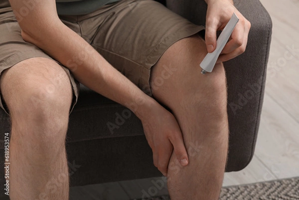 Fototapeta Man applying ointment from tube onto his knee indoors, closeup