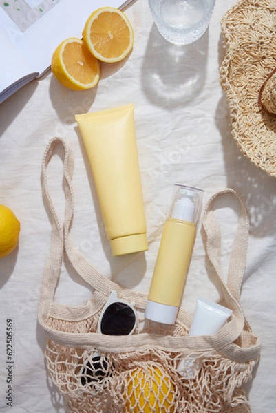 Fototapeta Summer concept of yellow cosmetic containers arranged with a hat, magazine, mesh bag with sunglasses and lemons. Mockup of skin care cosmetic extracted from Lemon (Citrus limon)