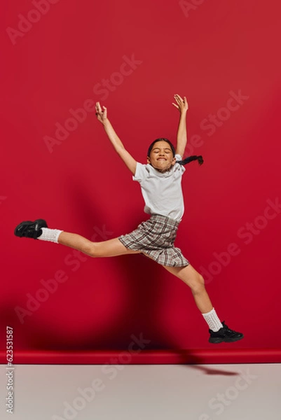 Fototapeta Excited and cheerful preteen girl in white t-shirt and stylish checkered skirt jumping and having fun while posing on red background, hairstyle and trendy accessories concept