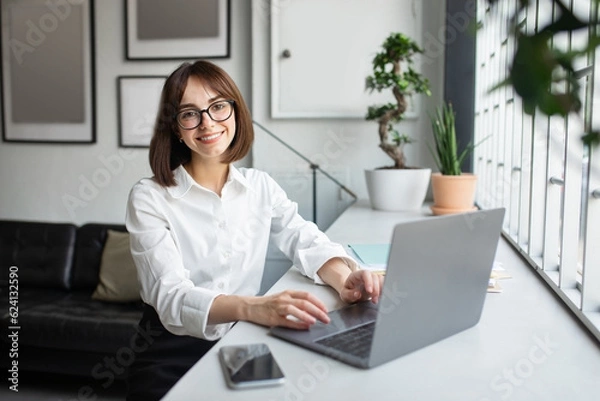 Fototapeta Successful freelancer. Happy lady sitting at desk with laptop and smiling at camera, working on computer in cozy office