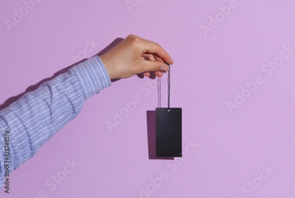 Fototapeta Man's hand in shirt holding blank black price tag on purple pastel background with shadow. Sale concept