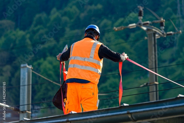 Fototapeta Male worker with high visibility clothing and helmet holding security belt on rooftop of platform of railway station at Swiss village of Flüelen. Photo taken May 22nd, 2023, Flüelen, Switzerland.