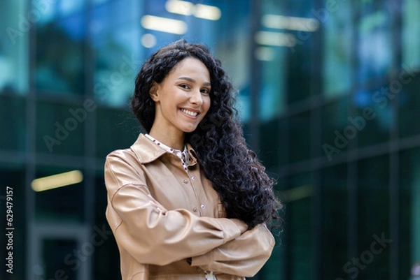 Fototapeta Portrait of happy and successful business woman, boss in shirt smiling and looking at camera inside office with crossed arms, Hispanic woman with curly hair outdoors.