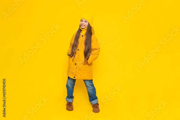 Fototapeta A young girl rejoices at the onset of autumn. Warm clothes for children. A child in an orange down jacket and hat on a yellow isolated background in the studio.