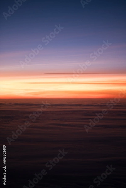 Fototapeta A radiant sunset, as seen from an airplane high above the clouds. The sky is a gradient of bluish purple and reddish orange. The light reflects off the tops of the clouds.