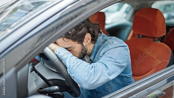 Fototapeta Young hispanic man sitting on car with head on steering wheel at street