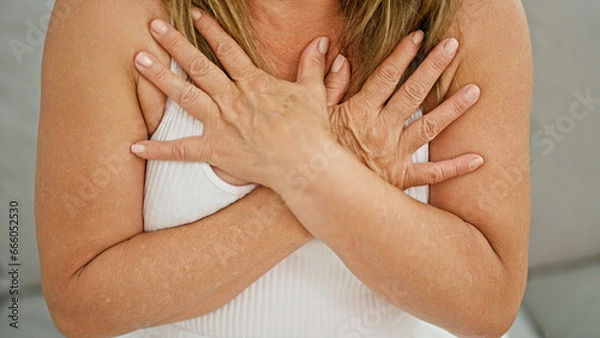 Fototapeta Middle age hispanic woman sitting on sofa with hands on chest at home