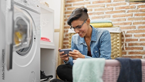 Fototapeta Young beautiful hispanic woman using smartphone waiting for washing machine at laundry room