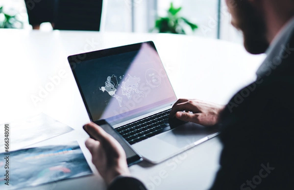 Fototapeta Businessman in suit sitting at desk in office typing on laptop while surfing mobile in workplace in company