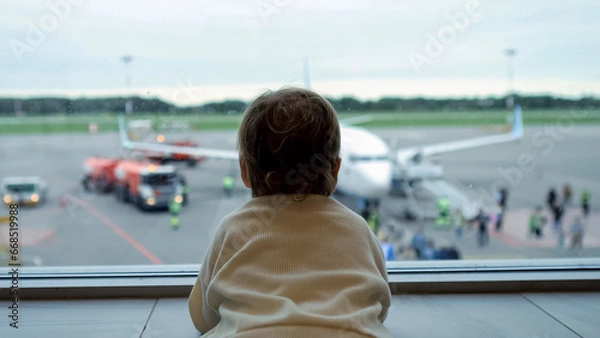 Fototapeta Toddler standing by large window looks at people getting aboard plane. Girl follows eyes for passengers preparing to embark on journey