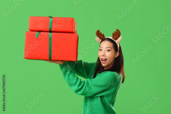 Fototapeta Young Asian woman in reindeer horns with Christmas gift boxes on green background