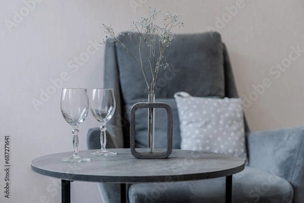 Obraz a round grey table. there are transparent glasses and a vase of flowers on the table. There is an armchair in the background.