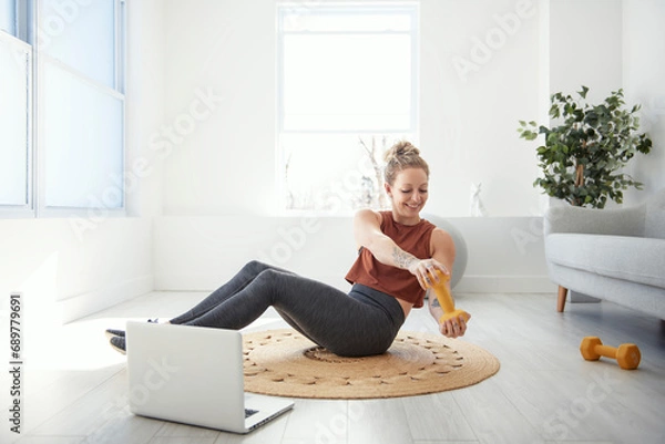 Fototapeta Cheerful active woman doing fitness at home, using laptop, watching online lessons.