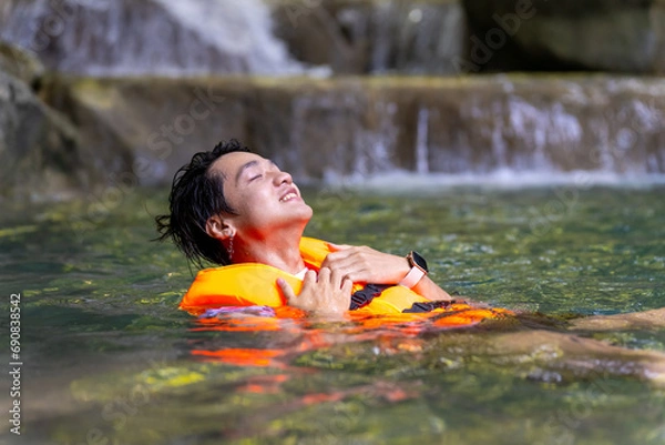 Fototapeta Young Asian man travel nature on summer holiday vacation. Happy generation z guy in life vest relax and enjoy outdoor lifestyle jumping and playing water at waterfall lagoon in forest mountain.