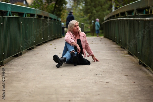 Fototapeta Woman blonde in her fifties with a pink denim jacket, blue jeans with a ripped black sweater and a black scarf, portrait on a bridge.