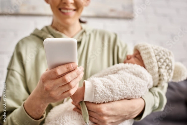 Fototapeta cheerful attractive woman in casual outfit looking at her mobile phone while holding her baby boy