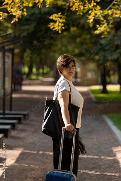 Fototapeta A middle-aged woman walks along the boulevard with a suitcase