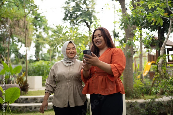 Fototapeta businesswoman holding and showing her phone to hijab coworker in happy expression