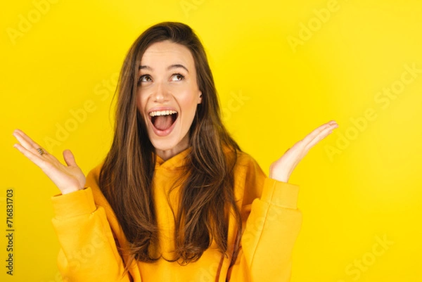 Fototapeta Optimistic woman raises palms from joy, happy to receive awesome present from someone, shouts loudly, dressed in yellow hoody, isolated on yellow background. Excited female yells, raised hand