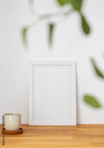 Fototapeta A white frame with a blank canvas against a light wall and on a wooden tabletop