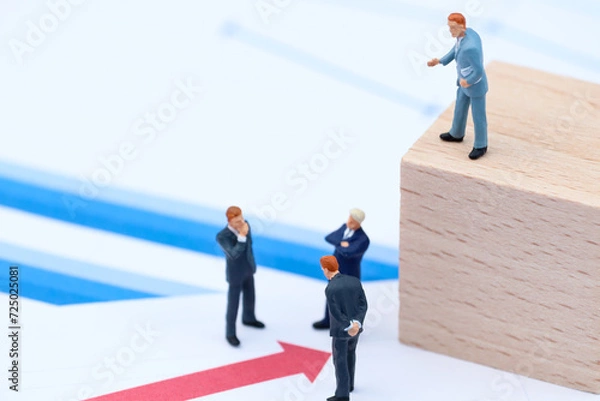 Fototapeta Miniature Business Leader Directing Team on Wooden Block Platform