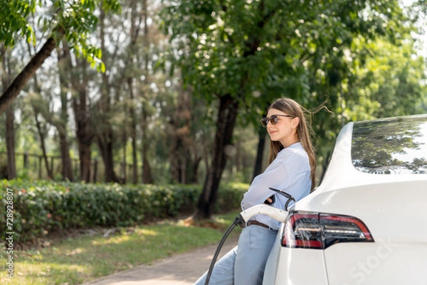 Fototapeta Young woman using smartphone online banking application to pay for electric car battery charging from EV charging station during vacation holiday road trip at national park or summer forest. Exalt