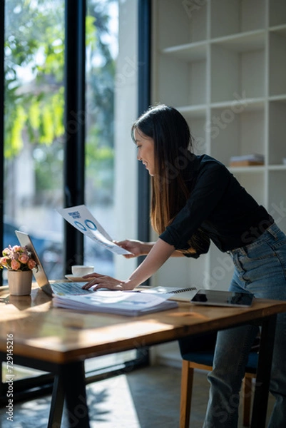 Fototapeta Cheerful businesswoman reviewing a financial report next to her laptop in a well-lit office space..