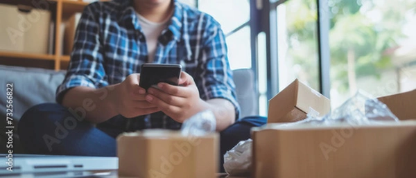 Fototapeta Man using smartphone amidst unpacking moving boxes