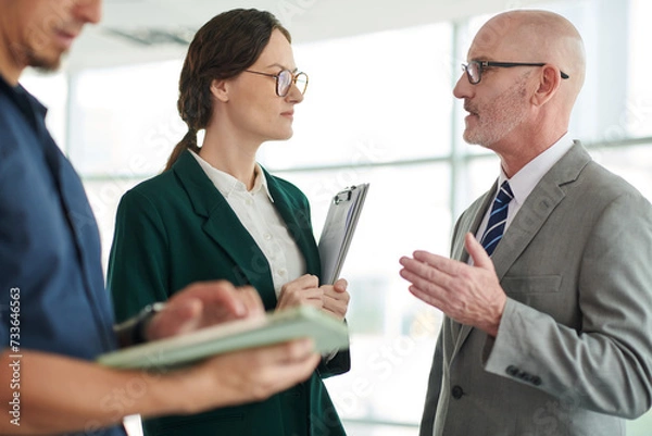 Fototapeta Confident male agent or broker in grey suit looking at young female colleague and explaining her contract terms and conditions