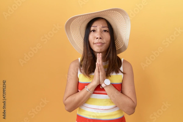 Fototapeta Middle age chinese woman wearing summer hat over yellow background begging and praying with hands together with hope expression on face very emotional and worried. begging.