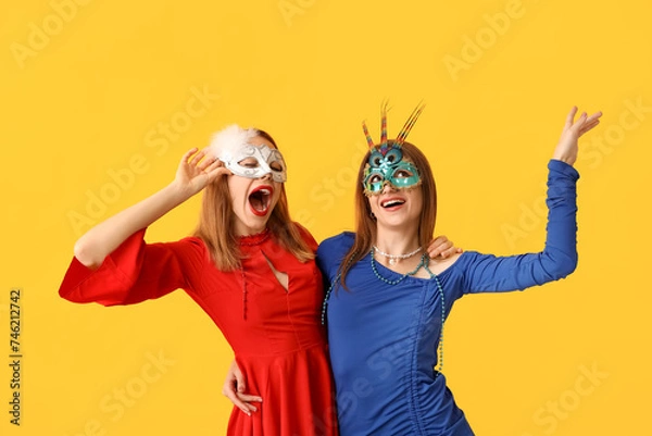 Fototapeta Shocked young women in carnival masks on yellow background