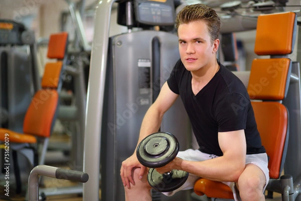 Fototapeta Young handsome man sits and raises dumbbell in left hand in fitness club